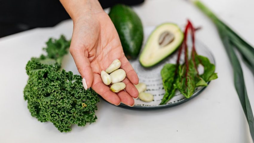 Close-up of a hand holding garlic cloves over a plate with fresh vegetables, including kale and avocado.