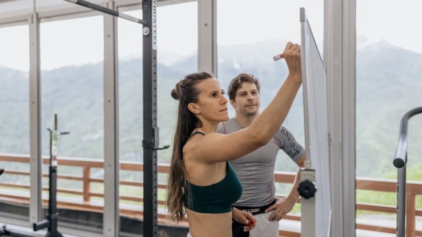 Female fitness coach writes on whiteboard as a male athlete watches in a gym.